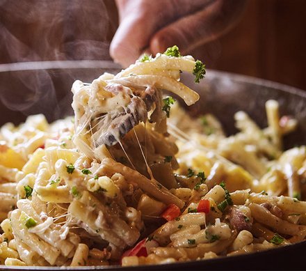 Traditionelle Älplermagronen mit Apfelmus Eine dampfende Pfanne mit Älplermagronen, die von einer Hand serviert werden