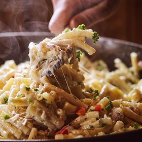 Traditionelle Älplermagronen mit Apfelmus Eine dampfende Pfanne mit Älplermagronen, die von einer Hand serviert werden