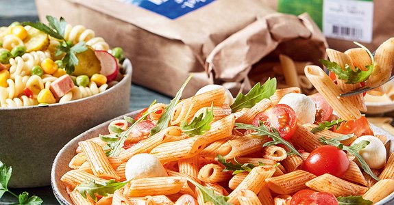 Frische Nudelsalate Zwei Schüsseln mit frischen Nudelsalaten, eine mit Fusilli, Erbsen, Mais und Gewürzgurken, die andere mit Penne, Kirschtomaten, Mozzarellakugeln und Rucola. Im Hintergrund sind ein Glas Gewürzgurken und eine Papiertüte mit Nudeln zu sehen.