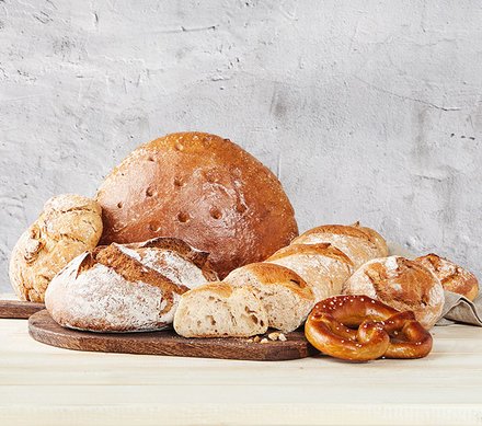 Brotvielfalt Eine Auswahl von verschiedenen Brotsorten, darunter ein großer Laib Brot, mehrere Baguettes, rundes Bauernbrot und eine Brezel, auf einem hellen Holztisch vor einer grauen Wand.