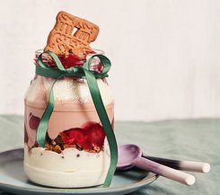 Spekulatius-Dessert im Glas Ein Glas-Dessert mit Spekulatius, Schichten aus Sahne, Kirschsoße und Schokoladenmousse, dekoriert mit einem grünen Band und Spekulatius-Keksen, serviert auf einem Teller mit zwei Löffeln.
