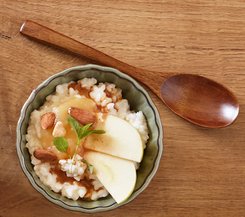 Milchreis mit Apfel und Mandeln Eine Schüssel Milchreis mit Apfelscheiben, Mandeln und Zimt, neben einem hölzernen Löffel auf einem Holztisch.