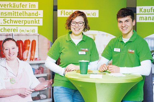 Zwei junge Auszubildende in grünen Polohemden stehen an einem grünen Stehtisch auf einer Messe. Hinter ihnen sind Werbeschilder für verschiedene Ausbildungsberufe.