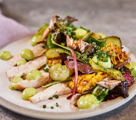 Gegrillter Hähnchensalat mit Avocadocreme Ein frischer Hähnchensalat mit gegrilltem Hähnchenbrustfilet, gemischtem Salat, gegrilltem Mais, Avocadocreme und Schnittlauch, angerichtet auf einem weißen Teller.