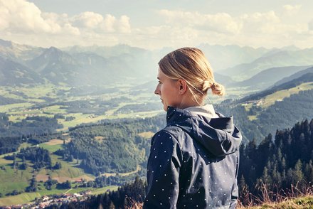 Amelie Feneberg auf Berggipfel mit Blick auf Landschaft Amelie Feneberg in einer blauen Jacke mit weißen Punkten steht auf einem Berg und blickt über eine malerische Landschaft mit grünen Tälern und bewaldeten Hügeln. Der Himmel ist bewölkt, und es herrscht klares Wetter.