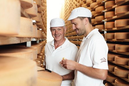 Käseproduktion im Lager Zwei Männer in weißen Arbeitskleidern und Mützen stehen in einem Käselager und überprüfen die Käseräder.