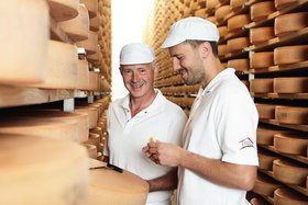 Zwei Männer in weißen Arbeitskleidern und Mützen stehen in einem Käselager und überprüfen die Käseräder.