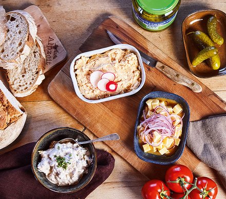 Allgäuer Tapas – Bayerische Brotzeit Brotzeit mit verschiedenen bayerischen Spezialitäten auf einem Holztisch, darunter Obazda, Radieschen, Essiggurken, Tomaten, Brot und Käse mit Zwiebeln