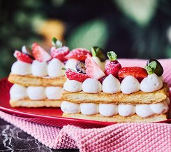 Blätterteigschnitte mit Tonkabohnencreme Blätterteigschnitten mit Tonkabohnencreme, dekoriert mit frischen Beeren und Minzblättern, serviert auf einem roten Teller mit einem rosa Stofftuch im Hintergrund.