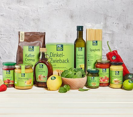 FE BIO-Produkte Eine Auswahl von FE BIO-Produkten, darunter Kaffee Crema, Dinkel-Zwieback, natives Olivenöl, Spaghetti, Erdbeerkonfitüre, Blütenhonig, Ahornsirup, Pesto Verde, Tomatensauce, mittelscharfer Senf, eine rote Paprika, eine Zitrone und frischer Spinat, auf einem hellen Holztisch vor einer grauen Wand.