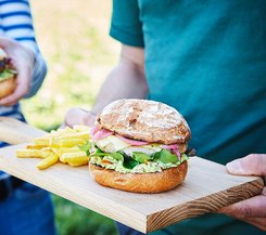 Bratwurstschnecken Burger Bratwurstschnecken Burger auf einem Holzbrett, serviert mit Pommes. Der Burger ist mit Salat, eingelegten Zwiebeln und Avocado belegt. Eine Person hält das Brett, während eine andere Person im Hintergrund einen ähnlichen Burger hält.