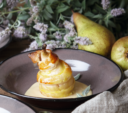 Birne im Blätterteig Birne im Blätterteigmantel auf einem Teller, serviert mit einer Creme und dekoriert mit frischen Kräutern, im Hintergrund sind weitere Birnen und Blumen zu sehen.
