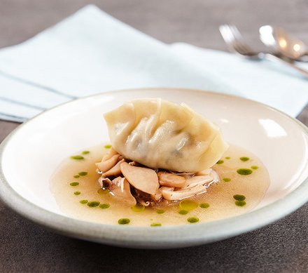 Gedämpfte Teigtasche mit Pilzen in Brühe Ein Teller mit einer gedämpften Teigtasche auf einem Bett aus Pilzen in Brühe, garniert mit grünen Öltröpfchen.