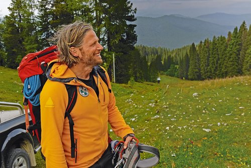 Ein Mann mit einer gelben Jacke und einem Rucksack steht auf einer blühenden Wiese mit Wald und Bergen im Hintergrund.