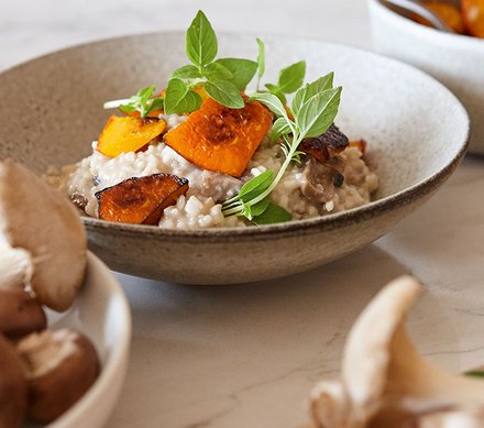 Cremiges Risotto mit geröstetem Kürbis Ein Teller cremiges Risotto, garniert mit geröstetem Kürbis und frischen Kräutern, im Hintergrund Pilze.