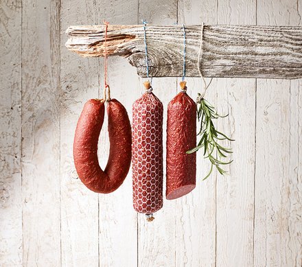 Wurstsorten an Holzbrett Drei verschiedene Wurstsorten hängen an einem rustikalen Holzbrett vor einer weiß gestrichenen Holzverkleidung. Eine der Würste ist ringförmig, während die anderen beiden gerade und mit Netzgewebe umhüllt sind. Ein Zweig Rosmarin hängt ebenfalls am Brett.