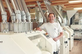 Ein Mann steht in einer modernen Mühle neben großen Maschinen und lächelt in die Kamera.
