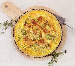 Quiche mit Speck und Spargelstreifen Eine herzhafte Quiche mit knusprigem Speck und Spargelstreifen auf einem runden Holzbrett, dekoriert mit frischen Kräutern.