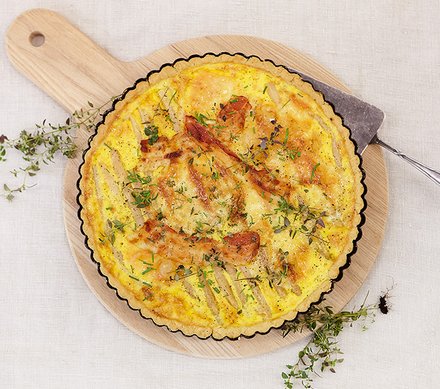 Quiche mit Speck und Spargelstreifen Eine herzhafte Quiche mit knusprigem Speck und Spargelstreifen auf einem runden Holzbrett, dekoriert mit frischen Kräutern.