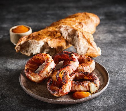 Bacon-Zwiebelringe mit Brot und Dip Goldbraune Bacon-Zwiebelringe auf einem Teller, serviert mit einer Scheibe knusprigem Brot und einer kleinen Schale Dip auf einem dunklen Hintergrund.