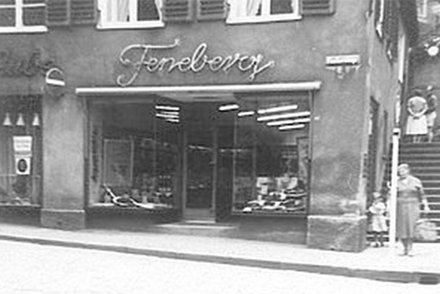 Historisches Feneberg-Geschäft Historische Schwarz-Weiß-Aufnahme eines Feneberg-Ladens. Der Laden hat große Schaufenster und ein handgeschriebenes Feneberg-Schild über dem Eingang. Zwei Personen, eine Frau und ein Kind, stehen draußen auf dem Gehweg.