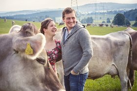 Bauer und Bäuerin stehen lächelnd inmitten ihrer Kühe auf einer grünen Wiese.