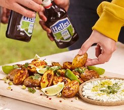 Gegrillte Kartoffeln mit Limetten und Kräutern Gegrillte Kartoffeln mit Limettenspalten und frischen Kräutern, serviert auf einem Holzbrett, neben einem Dip. Zwei Personen halten Bierflaschen.