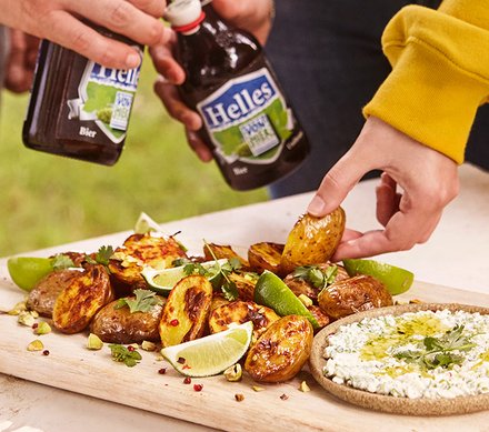 Gegrillte Kartoffeln mit Limetten und Kräutern Gegrillte Kartoffeln mit Limettenspalten und frischen Kräutern, serviert auf einem Holzbrett, neben einem Dip. Zwei Personen halten Bierflaschen.