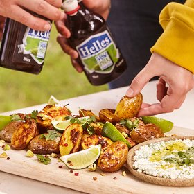Gegrillte Kartoffeln mit Limetten und Kräutern Gegrillte Kartoffeln mit Limettenspalten und frischen Kräutern, serviert auf einem Holzbrett, neben einem Dip. Zwei Personen halten Bierflaschen.