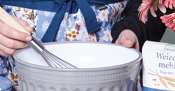 Backen mit frischen Zutaten Eine Person in einer blumigen Schürze hält einen Schneebesen in einer großen Schüssel, daneben stehen eine Packung Weizenmehl und ein Stück Butter. Eine andere Person hält einen Blumenstrauß.