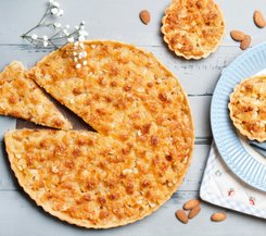 Apfeltartellets frisch gebacken Ein großer, goldbrauner Apfeltart mit einer geschnittenen Scheibe, umgeben von kleineren Apfeltartellets auf einem grauen Holztisch, dekoriert mit weißen Blüten und Mandeln.