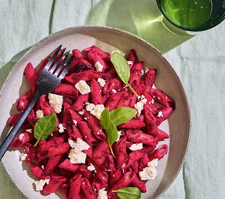Rote-Bete-Pasta mit Feta Ein Teller mit Rote-Bete-Pasta, garniert mit Feta-Käse und grünen Blättern, auf einem grünen Tuch serviert.