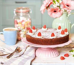 Dekorierte Torte mit Sahnehäubchen und Himbeeren Eine dekorierte Torte mit Sahnehäubchen und Himbeeren auf einer rosa Tortenplatte, im Hintergrund ein Kaffeebecher und ein Glas mit Süßigkeiten.