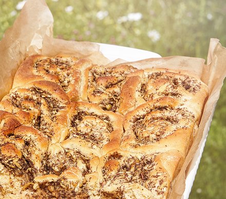 Apfel-Mohnschnecken frisch gebacken Eine frische, goldbraun gebackene Apfel-Mohnschnecke in einer mit Backpapier ausgelegten Form, draußen vor einer grünen Wiese.