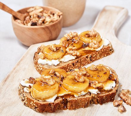 Honigbrot mit Bananen und Nüssen Zwei Scheiben Vollkornbrot mit Frischkäse, karamellisierten Bananen und gehackten Walnüssen auf einem Holzbrett, im Hintergrund eine Schale mit Nüssen