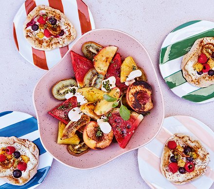 Bunter Obstsalat mit Fladenbrot Ein bunter Obstsalat mit Wassermelone, Kiwi, Ananas, Pfirsichen und einem Klecks Joghurt, serviert in einer pinken Schale, umgeben von kleinen Fladenbroten auf gestreiften Tellern.