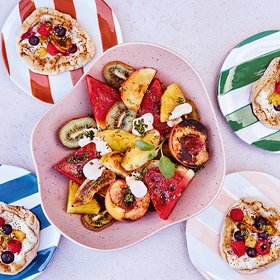 Bunter Obstsalat mit Fladenbrot Ein bunter Obstsalat mit Wassermelone, Kiwi, Ananas, Pfirsichen und einem Klecks Joghurt, serviert in einer pinken Schale, umgeben von kleinen Fladenbroten auf gestreiften Tellern.