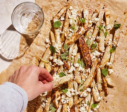 Gebackene Gemüse-Pommes mit Feta und Kräutern Eine Hand greift nach einer Portion gebackener Gemüse-Pommes, bestreut mit zerbröckeltem Feta und frischen Kräutern auf einem mit Backpapier ausgelegten Blech.