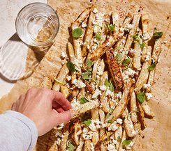 Gebackene Gemüse-Pommes mit Feta und Kräutern Eine Hand greift nach einer Portion gebackener Gemüse-Pommes, bestreut mit zerbröckeltem Feta und frischen Kräutern auf einem mit Backpapier ausgelegten Blech.