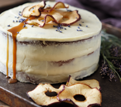 Lavendel-Apfel-Torte mit Karamell Eine Lavendel-Apfel-Torte mit weißem Frosting, dekoriert mit getrockneten Apfelscheiben und Lavendelblüten, mit Karamell, das an der Seite herunterläuft.