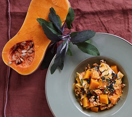 Butternut-Kürbis-Pasta mit frischen Kräutern Ein Teller mit Butternut-Kürbis-Pasta, garniert mit frischen Kräutern, neben einer halben Butternut-Kürbis.