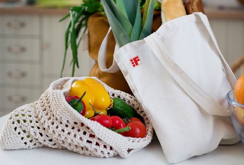 Eine Szene in einer Küche zeigt zwei wiederverwendbare Einkaufstaschen. Eine weiße Stofftasche mit einem roten "F" Logo ist gefüllt mit Lauch und Baguette. Daneben liegt eine gehäkelte Tasche mit gelber Paprika, Tomaten, Gurken und anderen frischen Gemüse.