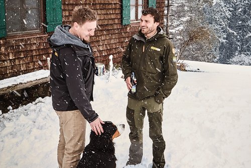 Zwei Männer, einer davon ein Ranger, stehen im Schnee und unterhalten sich. Ein Hund sitzt neben einem der Männer.