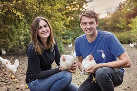 Ein junger Mann und eine junge Frau halten jeweils ein Huhn in den Händen und lächeln in die Kamera. Sie sitzen auf dem Boden einer Freilandhühnerhaltung.
