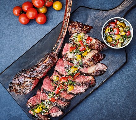 Gegrilltes Tomahawk-Steak mit Tomaten-Salsa Ein perfekt gegrilltes Tomahawk-Steak, in Scheiben geschnitten und mit einer frischen Tomaten-Salsa garniert. Daneben liegen gelbe und rote Kirschtomaten.