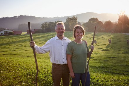 Bauernpaar in der Abendsonne Ein Mann und eine Frau stehen mit Wanderstöcken auf einer grünen Wiese bei Sonnenuntergang, mit Hügeln und Bäumen im Hintergrund.