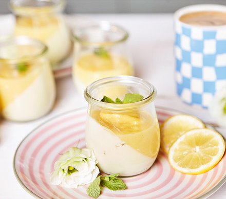 Zitronencreme-Dessert im Glas Kleine Gläser mit Zitronencreme-Dessert, dekoriert mit Minzblättern, auf einem gestreiften Teller mit Zitronenscheiben und einer Blüte.