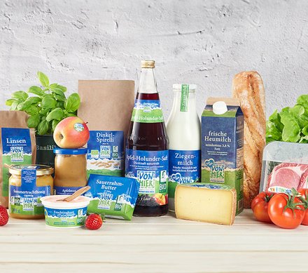 Regionale und Biologische Produkte Eine Auswahl von regionalen und biologischen Produkten, darunter Apfel-Holundersaft, Ziegenmilch, frische Heumilch, Linsen, Dinkel-Spirelli, Sommertrachthonig, Sauerrrahmbutter, verschiedene Käsesorten, Tomaten, ein Baguette, eine Basilikumpflanze, ein Apfel und Erdbeeren, auf einem hellen Holztisch vor einer grauen Wand.